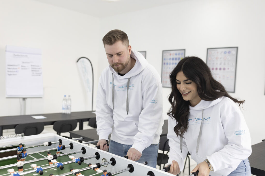 Ein Foto des Teams von Fast Drive Wuppertal, bestehend aus einer Frau und einem Mann, die vor dem Theorieunterricht entspannen und Spaß haben. Sie spielen zusammen am Kicker-Tisch, was Teil des modernen und abwechslungsreichen Ansatzes der Fahrschule ist. Dies fördert Teambuilding, Interaktion und schafft Erfolgsmomente, wenn sie gewinnen. Diese Abbildung vermittelt das engagierte und positive Arbeitsumfeld sowie den Fokus auf Spaß und Zusammenarbeit im Team.