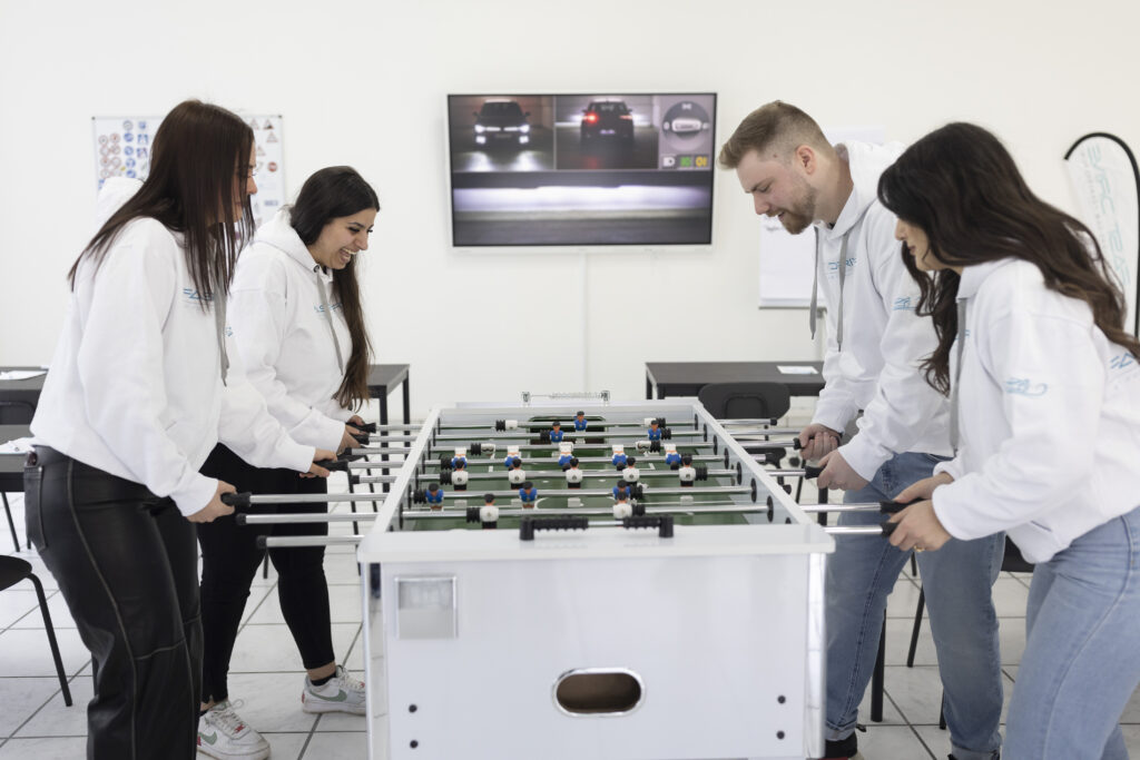 Ein Foto des Teams von Fast Drive Wuppertal, bestehend aus drei Frauen und einem Mann, die vor dem Theorieunterricht entspannen und Spaß haben. Sie spielen zusammen am Kicker-Tisch, was Teil des modernen und abwechslungsreichen Ansatzes der Fahrschule ist. Dies fördert Teambuilding, Interaktion und schafft Erfolgsmomente, wenn sie gewinnen. Diese Abbildung vermittelt das engagierte und positive Arbeitsumfeld sowie den Fokus auf Spaß und Zusammenarbeit im Team.