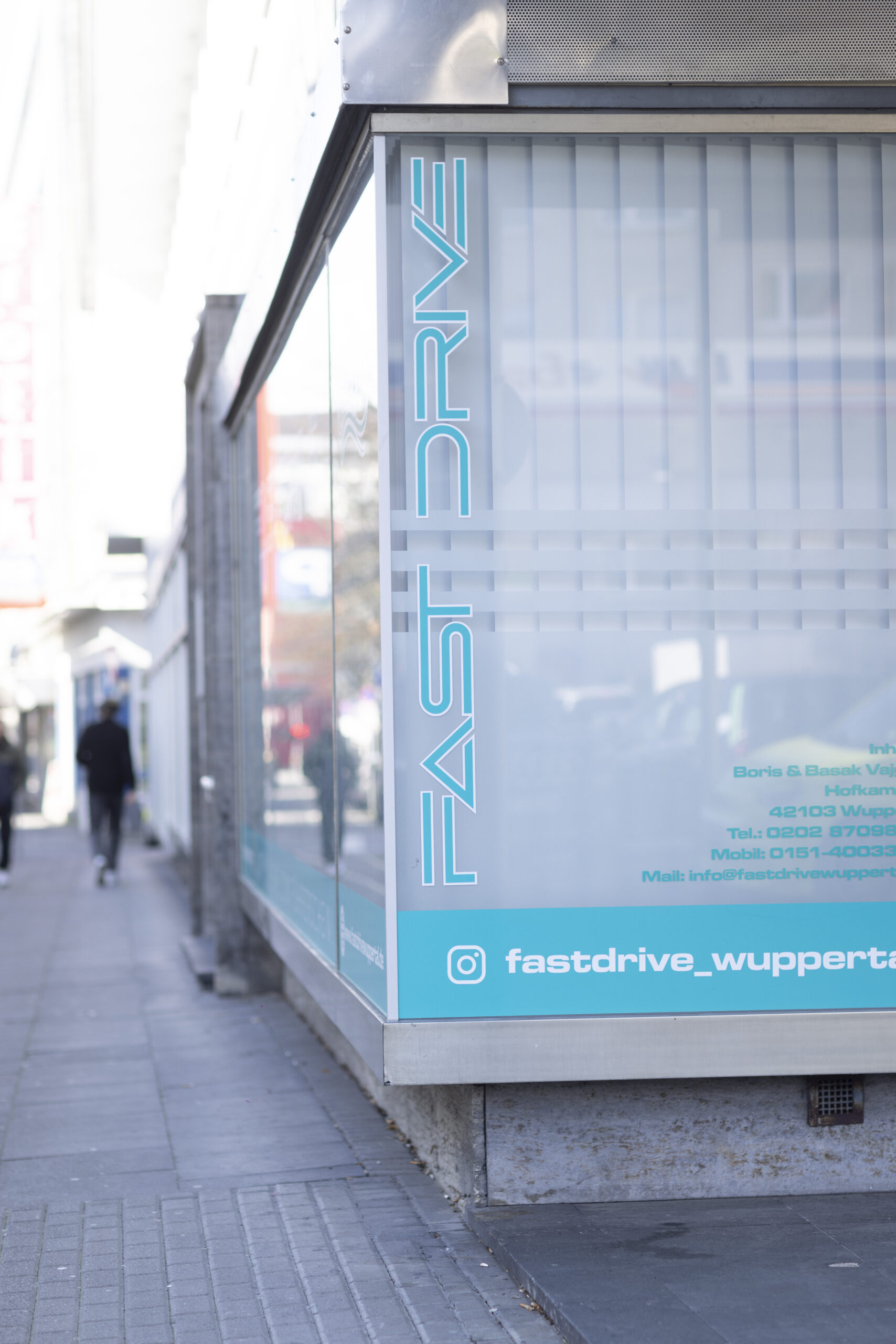 Die Außenfassade der Fahrschule in Wuppertal Elberfeld, befindlich in der Hofkamp 35, 42103 Wuppertal. Gut sichtbar ist die Aufschrift "Fast Drive" mit dem Slogan "Mit Vollgas zum Führerschein". Unterhalb sind Symbole für die Social Media Kanäle der Fahrschule zu erkennen. Diese Abbildung repräsentiert die Präsenz und Erreichbarkeit der Fahrschule sowie ihre moderne Marketingstrategie.