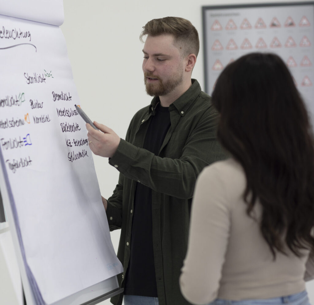 Ein Fahrlehrer hält einen Theorieunterricht, während er ein spezifisches Thema am Whiteboard erklärt. Eine Fahrschülerin hört aufmerksam zu, während sie persönliche Betreuung und eine gezielte Vermittlung von Lerninhalten erhält. Dies veranschaulicht den professionellen Theorieunterricht bei Fast Drive Wuppertal, der Fahrschule in Wuppertal Elberfeld.