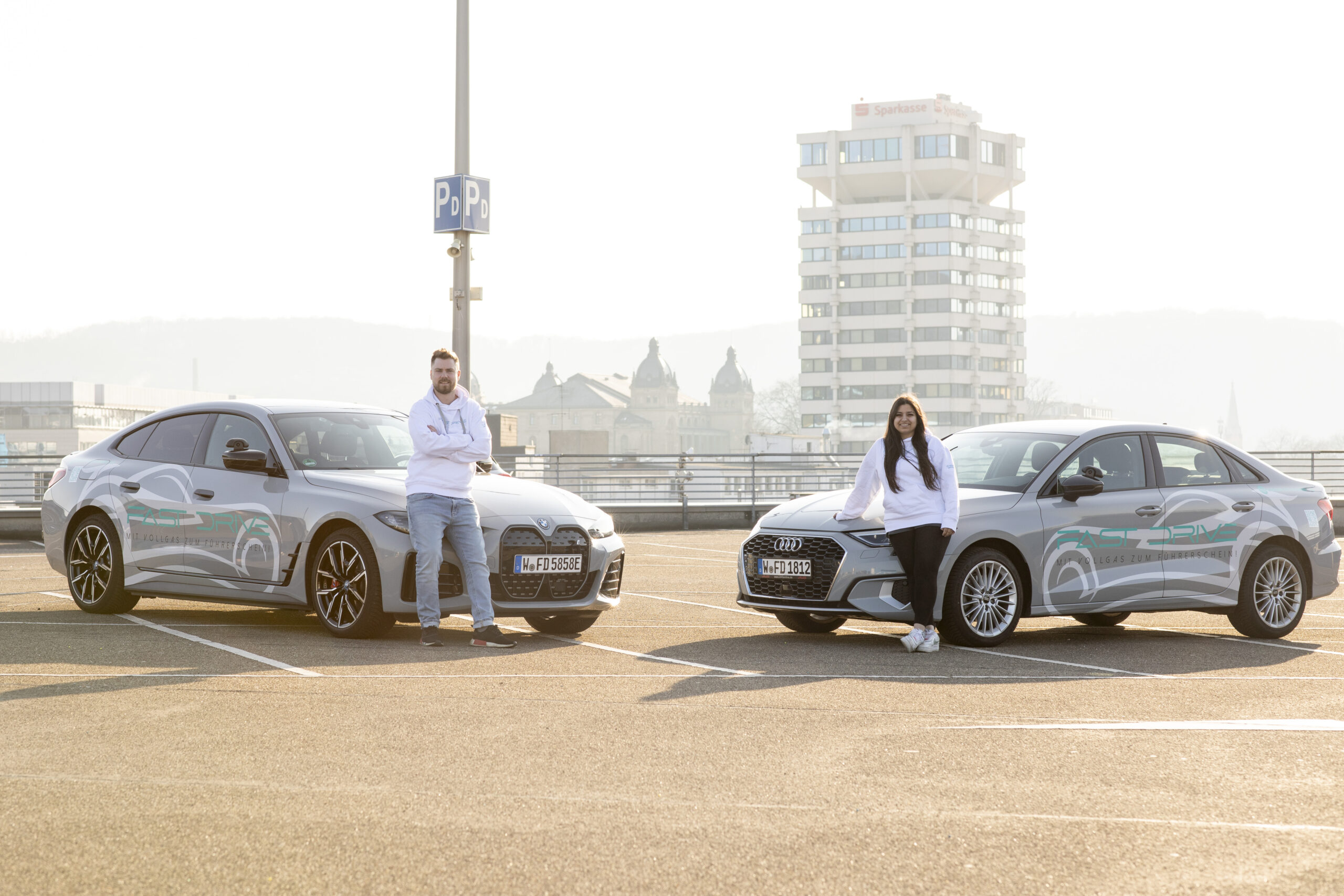 Zu sehen sind beide Fahrschulautos mit der markanten Fast Drive Folierung, die Sportlichkeit und Eleganz verkörpern. Daneben stehen beide Fahrschullehrer, die gleichzeitig die Besitzer sind. Sie repräsentieren die beiden Teams der Fahrschule: Team Beast und Team Eleganz. Diese Abbildung vermittelt den Charakter und die Leitung sowie ihre Markenidentität.