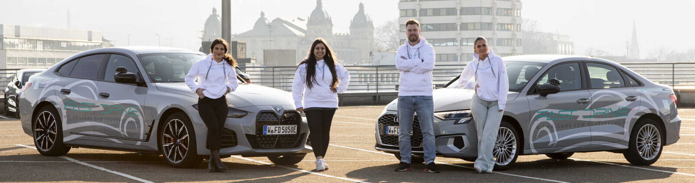 Das Fast Drive Team für die Fahrschule Wuppertal Barmen. Zu sehen sind die zwei Fahrschulbesitzer und Fahrlehrer in der Mitte anlehnend an die Fahrschulautos (BMW i4 Gran Coupe, links und AUDI A3 Limousine, rechts) und außen – jeweils links und rechts – die zwei Servicemitarbeiterinnen, die sich ebenfalls an die Autos anlehnen. Gemeinsam posieren Sie auf dem Parkdeck.